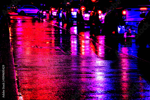 A group of people are standing on a wet street at night. The street is illuminated by the lights of cars and police cars. The scene is dark and moody