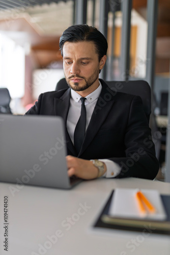 A man in a suit is sitting at a desk with a laptop open in front of him. He is wearing a tie and he is focused on his work