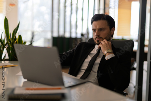 A man in a suit is sitting at a desk with a laptop open in front of him. He is wearing a tie and he is focused on his work