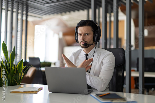 A man wearing a headset is sitting at a desk with a laptop in front of him. He is making a hand gesture, possibly indicating frustration or annoyance
