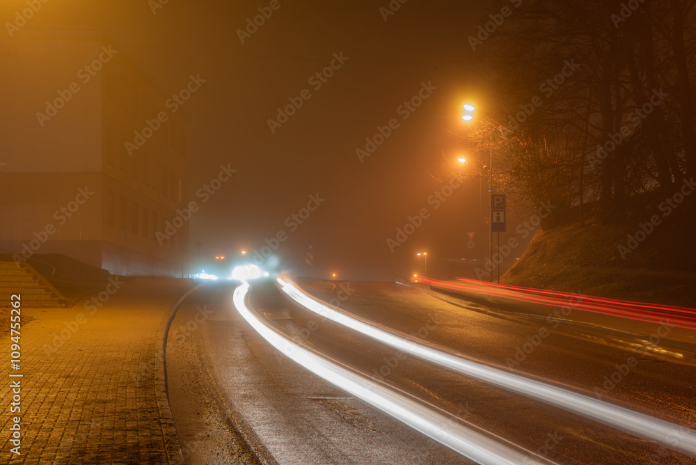 A foggy urban street at night with car light trails, glowing streetlights, a building, and trees creating a mysterious and atmospheric scene. A foggy urban street at night with car light trails, glowing streetlights, a building, and trees creating a mysterious and atmospheric scene.