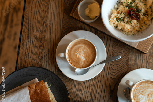 Breakfast setup featuring a creamy cappuccino with intricate latte art served in a white ceramic cup. Table showcases a delicious combination of fresh scrambled eggs garnished with herbs, tomatoes