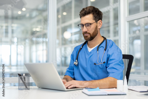 Man doctor in blue scrubs works on laptop in bright, modern medical office. Doctor wearing stethoscope, using technology for research and communication. Concept of healthcare, medicine, professional.