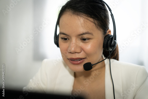 Female operator   wearing a headset is talking on the phone. She is wearing a white jacket and is sitting at a desk