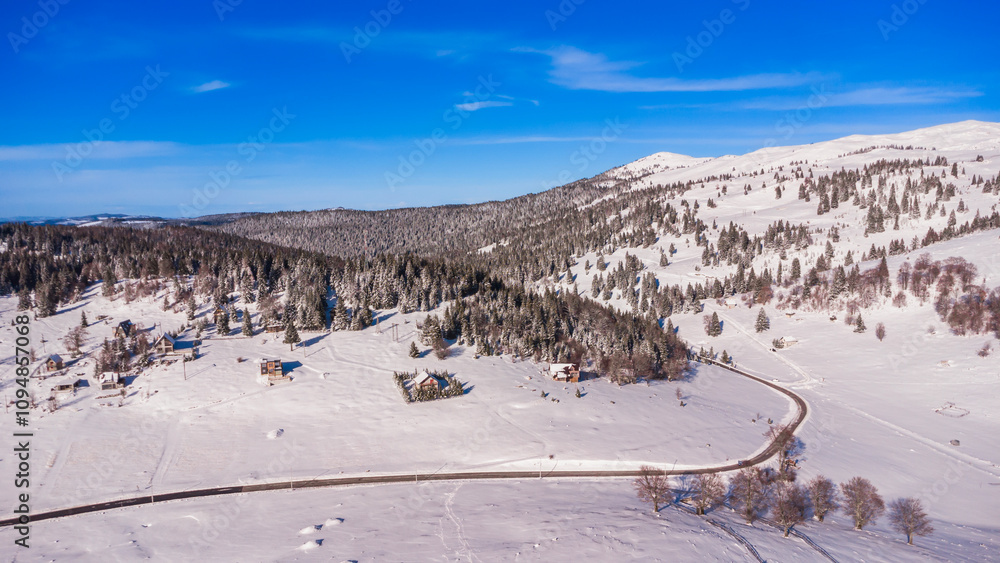 Fototapeta premium An aerial view of a peaceful snow-covered valley with scattered houses, surrounded by dense evergreen forests and rolling hills. The winding road cuts through the pristine winter landscape 