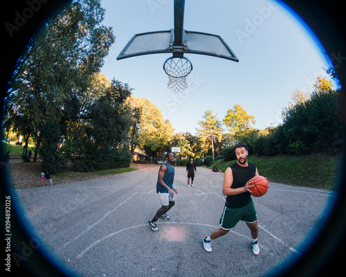 Two basketball players engage in a fierce contest for possession on an outdoor court surrounded by lush trees, showcasing competitive energy and athleticism under soft natural light.