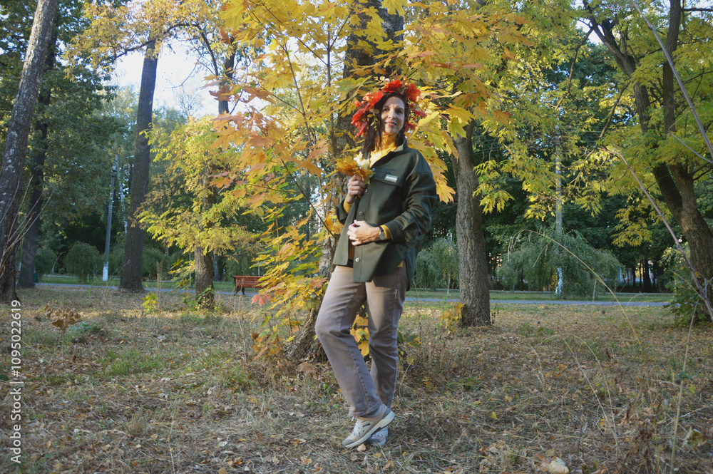 Happy smiling young girl, woman in wreath of autumn leaves against background of trees with yellow foliage. Autumn concept. Happy smiling young girl, woman in wreath of autumn leaves against background of trees with yellow foliage. Autumn concept.