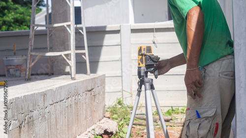 Builder uses laser to measure level of concrete house structure, worker works outdoors in construction site.