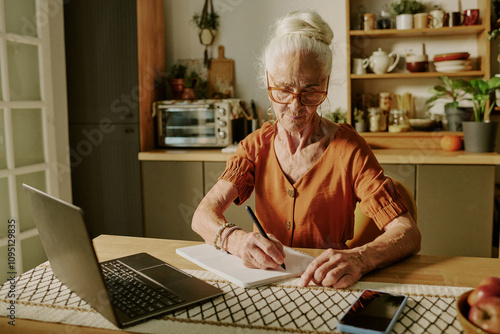 Portrait of senior woman writing on paper while sitting at desk with laptop in cozy kitchen, surrounded by kitchen utensils and decor photo