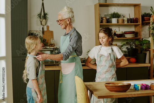 Obraz na plátně Elderly woman in apron standing in a modern kitchen engaging with two children while preparing food