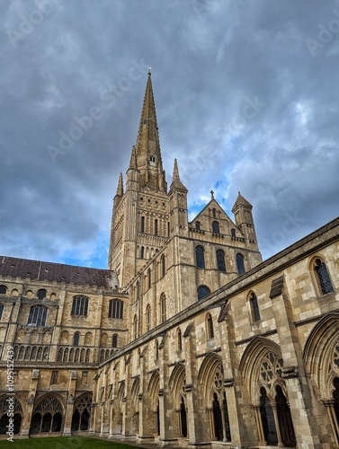 A breathtaking view of Norwich Cathedral, showcasing its impressive Gothic architecture, soaring spires, and intricate stonework. The dramatic sky adds to the grandeur of this historic landmark.