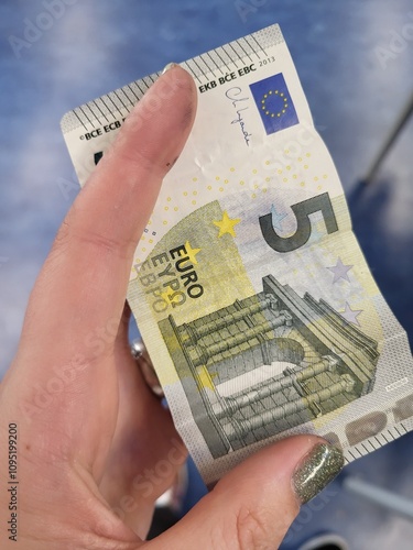 A close-up photo of a five-euro banknote being held in a hand. The banknote shows the iconic European architecture and the signature of the European Central Bank President.
