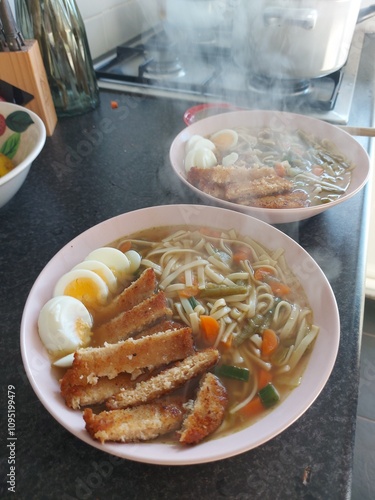 A steaming bowl of homemade ramen with noodles, vegetables, soft-boiled eggs, and crispy fried tofu. The dish is presented on a pink plate and looks both comforting and delicious.