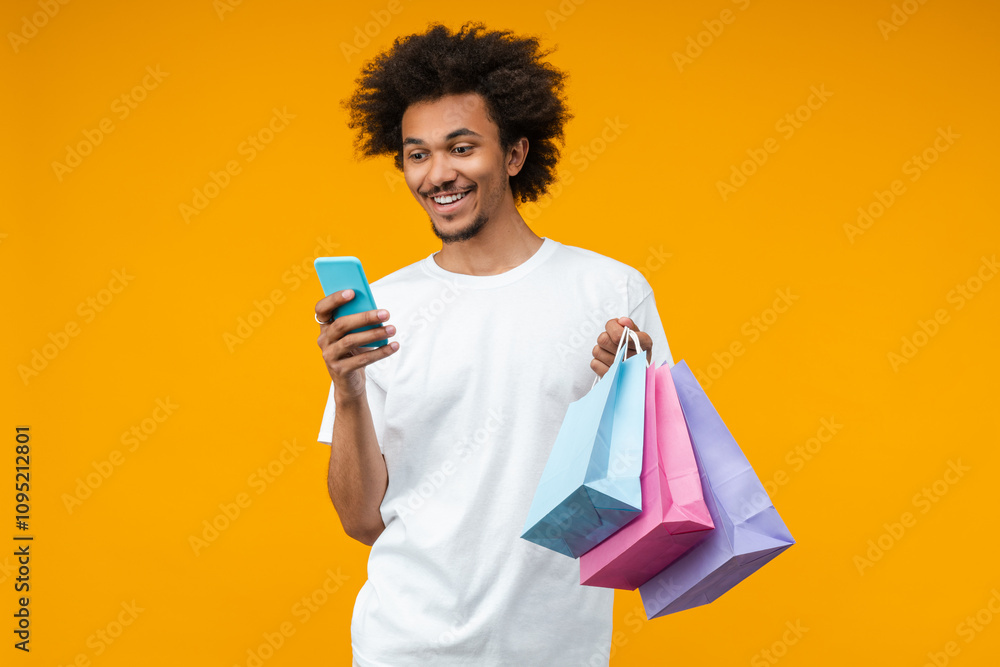 Studio portrait of handsome smiling young man customer with a bunch of shopping bags is making online purchases using his mobile phone, isolated over bright colored orange yellow background Studio portrait of handsome smiling young man customer with a bunch of shopping bags is making online purchases using his mobile phone, isolated over bright colored orange yellow background