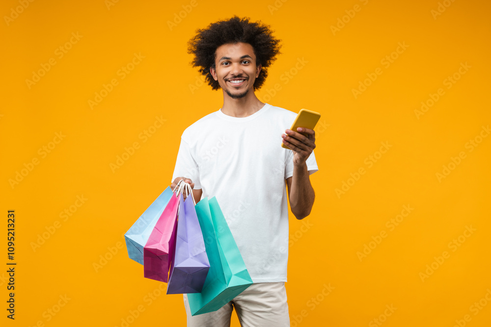 Attractive smiling African American man customer posing in studio with a bunch of shopping bags and mobile phone in hands isolated over orange yellow background Attractive smiling African American man customer posing in studio with a bunch of shopping bags and mobile phone in hands isolated over orange yellow background