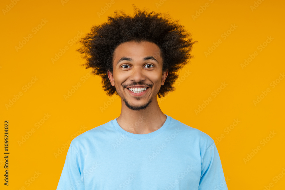Close up portrait of young African American man in basic blue t-shirt posing in studio, isolated over yellow background Close up portrait of young African American man in basic blue t-shirt posing in studio, isolated over yellow background