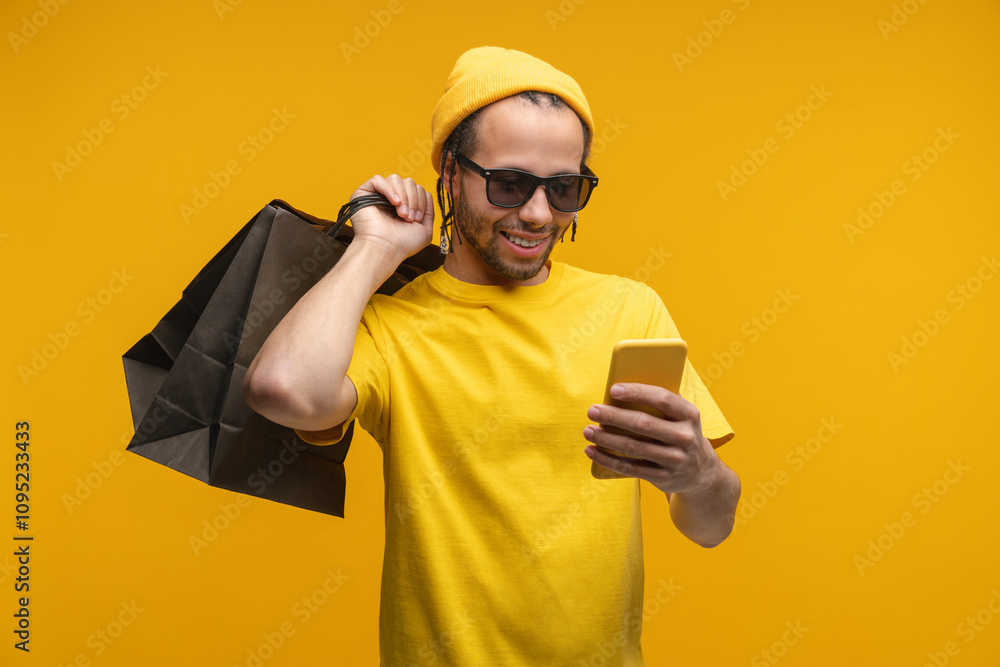 Studio portrait of young handsome smiling African American man browsing on his smartphone while standing over yellow background holding a bunch of shopping bags Studio portrait of young handsome smiling African American man browsing on his smartphone while standing over yellow background holding a bunch of shopping bags