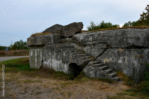 View of a damaged and destroyed bunker or bomb shelter from World War II located next to a dirt path, some river or lake and a vast area of lush meadows, forests, and moors seen near sunset in Poland photo