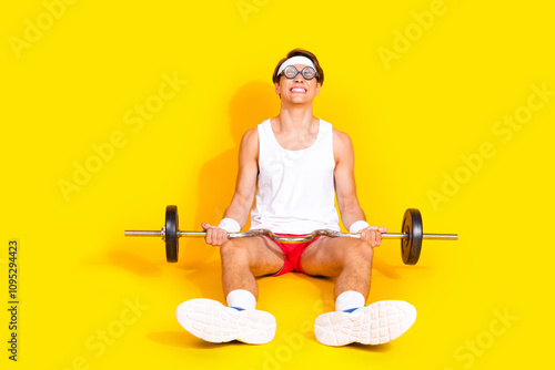 Energetic young man in activewear seated on vibrant yellow background with barbell, embodying healthy lifestyle and fitness motivation