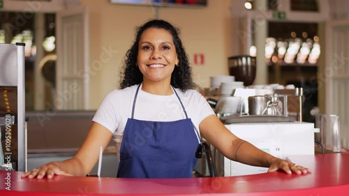 Confident Latina shop owner smiling behind store counter in apron. Proud small businesswoman posing at retail counter