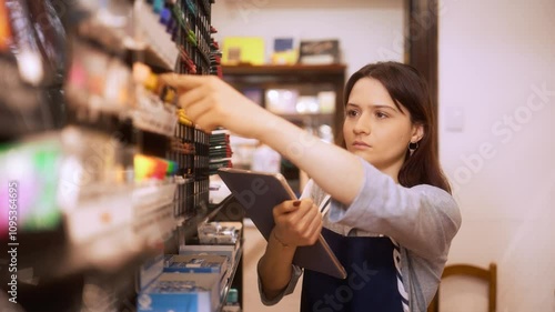 Focused store employee checking inventory and organizing products on the shelf. Female manager carefully examining items while holding a digital tablet in a retail store