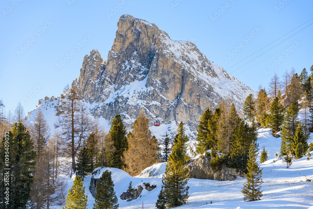 Majestic snow-capped rocky peak overlooking a ski resort in the Alps on a clear winter day. A red aerial tramway going up is visible against the mountain face. Majestic snow-capped rocky peak overlooking a ski resort in the Alps on a clear winter day. A red aerial tramway going up is visible against the mountain face.