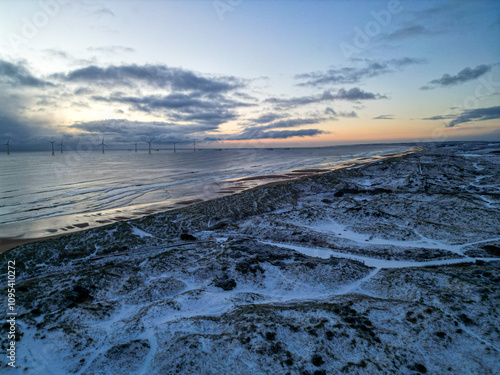 Aerial view over snow covered sand dunes on beach in Aberdeenshire, Scotland. Wind turbines and boats sit out in the North Sea during sunrise. 