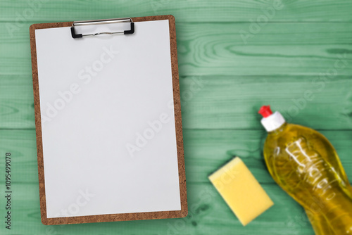 Clipboard checklist Blank, bottle of detergent, kitchen sponge on green table. Sheet of paper on Clip board with Place for text, messages, list, flat lay. Planning housekeeping spring cleaning.