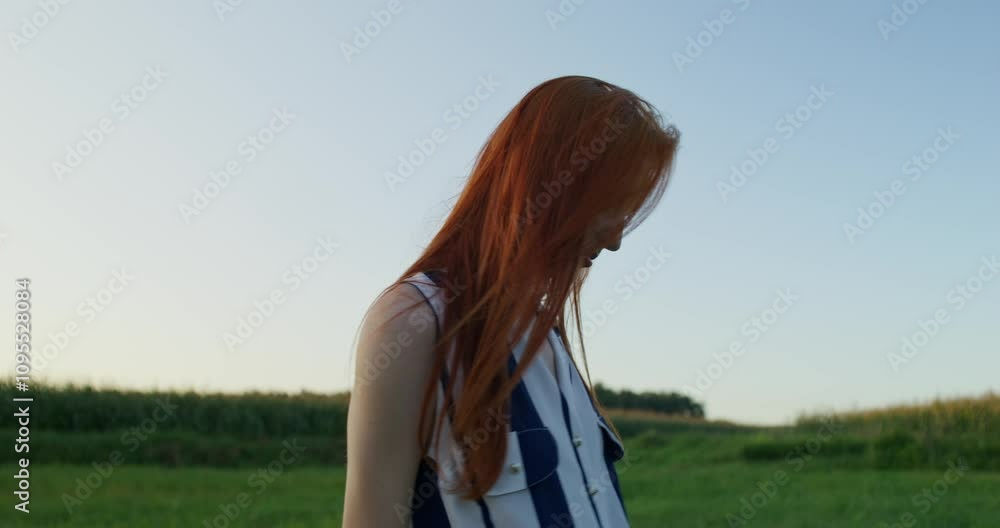 A red-haired woman smiles at the camera while walking through a field on a summer evening. A red-haired woman smiles at the camera while walking through a field on a summer evening.