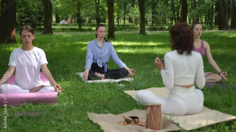 A meditation teacher in white guides a group session in a tranquil park using a rainstick to create soothing sounds. Two participants sit cross-legged on mats, focusing on their breathing and A meditation teacher in white guides a group session in a tranquil park using a rainstick to create soothing sounds. Two participants sit cross-legged on mats, focusing on their breathing and