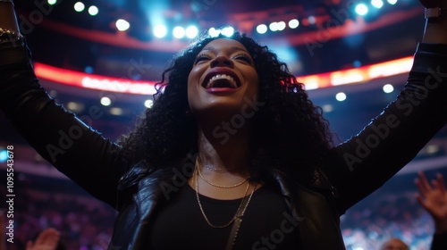 A joyful woman with curly hair raises her arms in celebration while enjoying a concert in an energetic arena. The crowd around her is engaged in the lively atmosphere, sharing the moment. photo