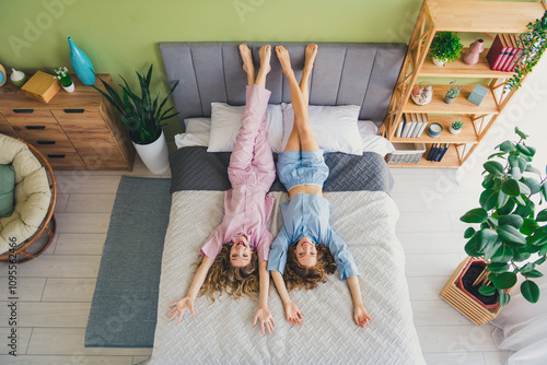 Full size photo of two young women have fun lying bed raise legs upside down dressed sleepwear outfit wake up cozy bedroom home interior