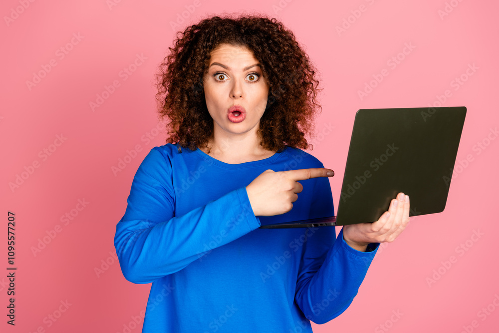 Surprised young woman pointing at laptop against a pink background wearing a casual blue sweatshirt Surprised young woman pointing at laptop against a pink background wearing a casual blue sweatshirt