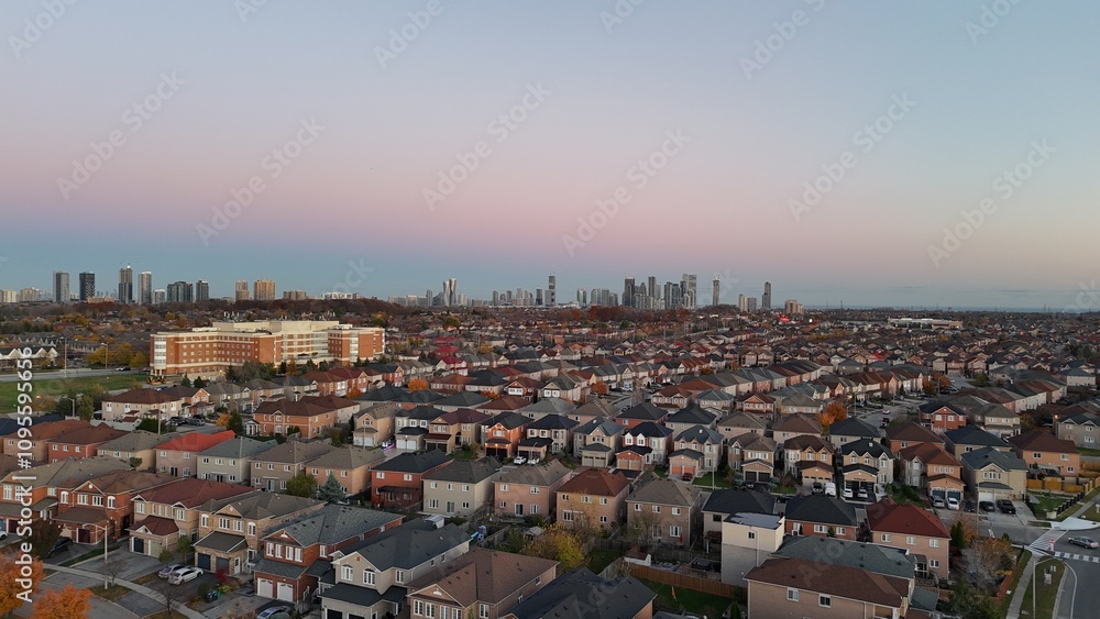 Obraz premium A scenic aerial view of a suburban neighborhood during sunset, with rows of colorful houses leading to a distant city skyline under a gradient sky transitioning from pink to blue. A vibrant blend of u