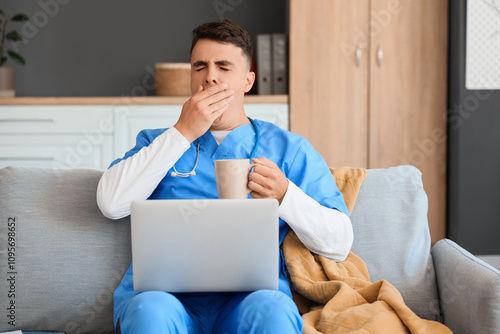 Male medical student with cup of coffee and laptop yawing on sofa at home photo