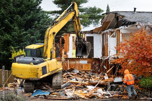 Bild auf Leinwand Large yellow excavator starting demolition on a house, workman spraying water to