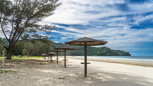 A row of sun umbrellas on the sandy beach. Green grass, trees on the lawn. The waves of the turquoise ocean are foaming. A hill in the distance against the blue sky and clouds. Malaysia. Borneo. 