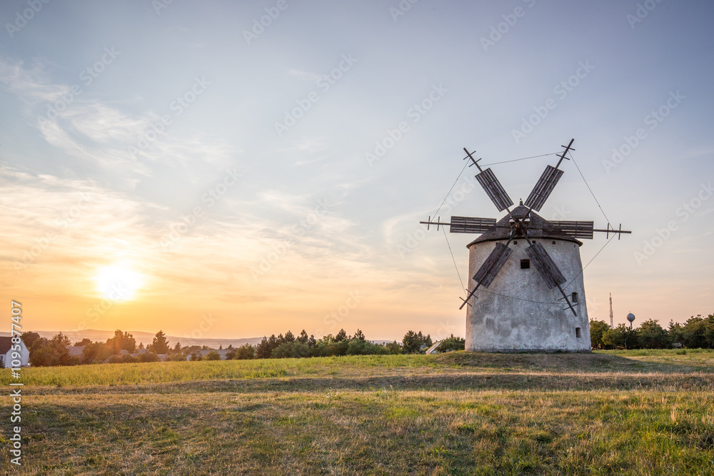 Windmill with wooden wings in a landscape setting. Fields, meadows and flowers appear in the sunset. Landscape shot in nature with a mill. historically untypical for the Balaton region in Hungary Windmill with wooden wings in a landscape setting. Fields, meadows and flowers appear in the sunset. Landscape shot in nature with a mill. historically untypical for the Balaton region in Hungary