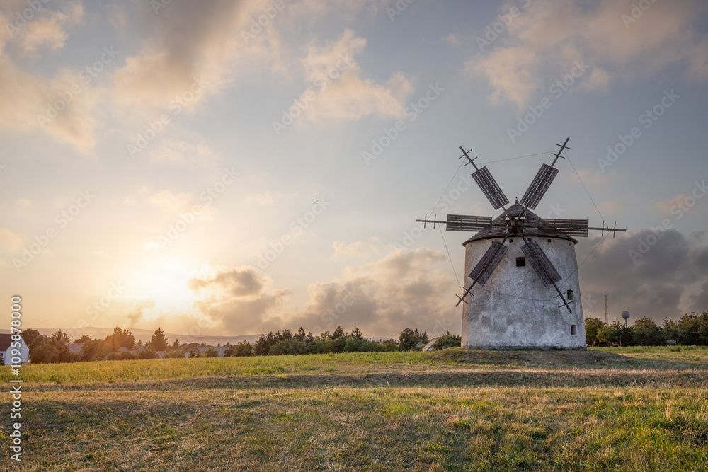 Windmill with wooden wings in a landscape setting. Fields, meadows and flowers appear in the sunset. Landscape shot in nature with a mill. historically untypical for the Balaton region in Hungary Windmill with wooden wings in a landscape setting. Fields, meadows and flowers appear in the sunset. Landscape shot in nature with a mill. historically untypical for the Balaton region in Hungary