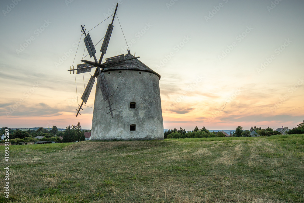 Windmill with wooden wings in a landscape setting. Fields, meadows and flowers appear in the sunset. Landscape shot in nature with a mill. historically untypical for the Balaton region in Hungary Windmill with wooden wings in a landscape setting. Fields, meadows and flowers appear in the sunset. Landscape shot in nature with a mill. historically untypical for the Balaton region in Hungary