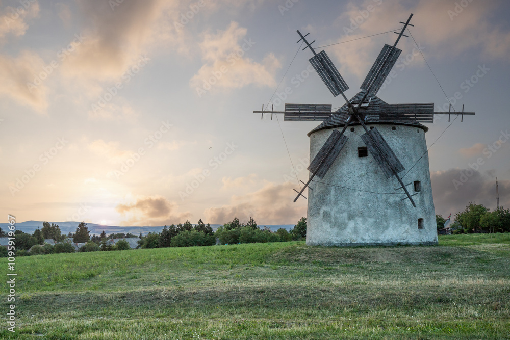 Windmill with wooden wings in a landscape setting. Fields, meadows and flowers appear in the sunset. Landscape shot in nature with a mill. historically untypical for the Balaton region in Hungary Windmill with wooden wings in a landscape setting. Fields, meadows and flowers appear in the sunset. Landscape shot in nature with a mill. historically untypical for the Balaton region in Hungary