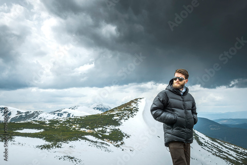 Young man with a beard standing in snowy mountain area wearing cold weather accessories, warm down jacket against a scenic backdrop of snowy mountains and snow-laden clouds.