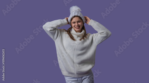 Cheerful young woman in white clothing and knitted hat smiling holding hands behind head, isolated on purple studio background. Middle shot photo