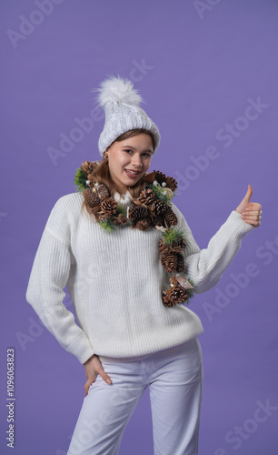 Cheerful young woman in white clothing and knitted hat shows like thumb up gesture, isolated on purple studio background. photo