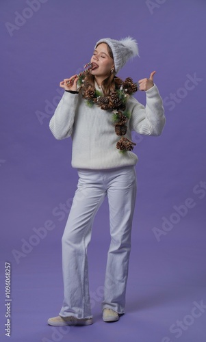 Cheerful young woman in white clothing and knitted hat with candy cane showing thumbs up, isolated on purple studio background. Full body. photo