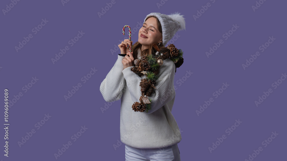 Cheerful young woman in white clothing and knitted hat posing with closed eyes holding candy cane, isolated on purple studio background. Middle shot. Cheerful young woman in white clothing and knitted hat posing with closed eyes holding candy cane, isolated on purple studio background. Middle shot.