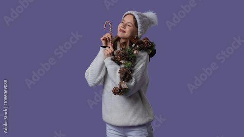 Cheerful young woman in white clothing and knitted hat posing with closed eyes holding candy cane, isolated on purple studio background. Middle shot. photo