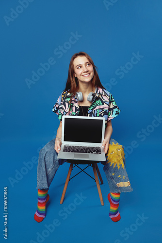 Cute student lady with smiling and demonstrating new modern laptop with empty screen, isolated on blue background. Excited girl with headphones sitting on chair and holding laptop while looking away.