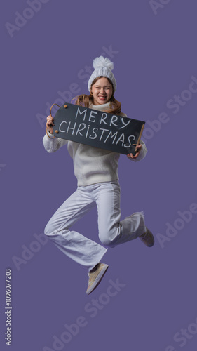 Cheerful young woman in white clothing and knitted hat jumps holding chalkboard with merry christmas text, isolated on purple studio background. Full body. photo