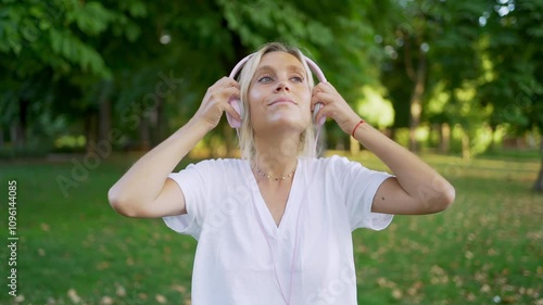 Young woman enjoys music while putting on headphones in a sunny park, surrounded by nature and greenery